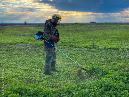 A man tightly grips a hand-held gasoline scythe and with quick and precise movements skillfully cuts a clover log at sunset. He is wearing a hazmat suit and a helmet that protects his face.