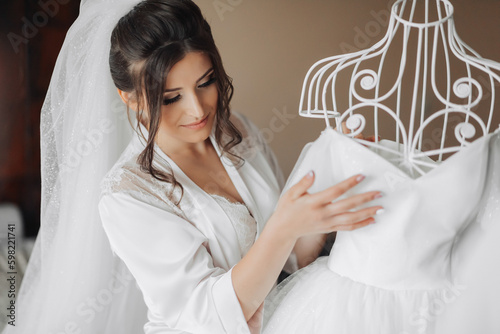 Brunette bride in a robe  posing for the camera  looking at her wedding dress. Elegant hairstyle. Nice makeup. Voluminous veil. Morning of the bride