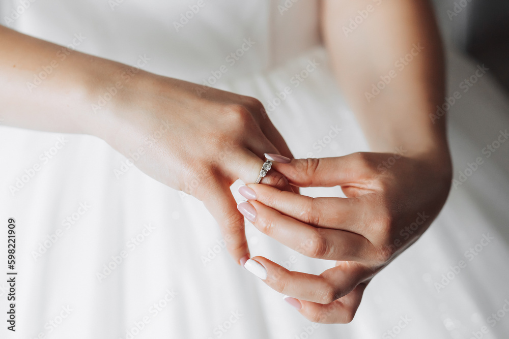 A brunette bride in a white dress puts on her silver wedding ring. French manicure. Open shoulders. Beautiful hands. Long veil. Morning of the bride. Details