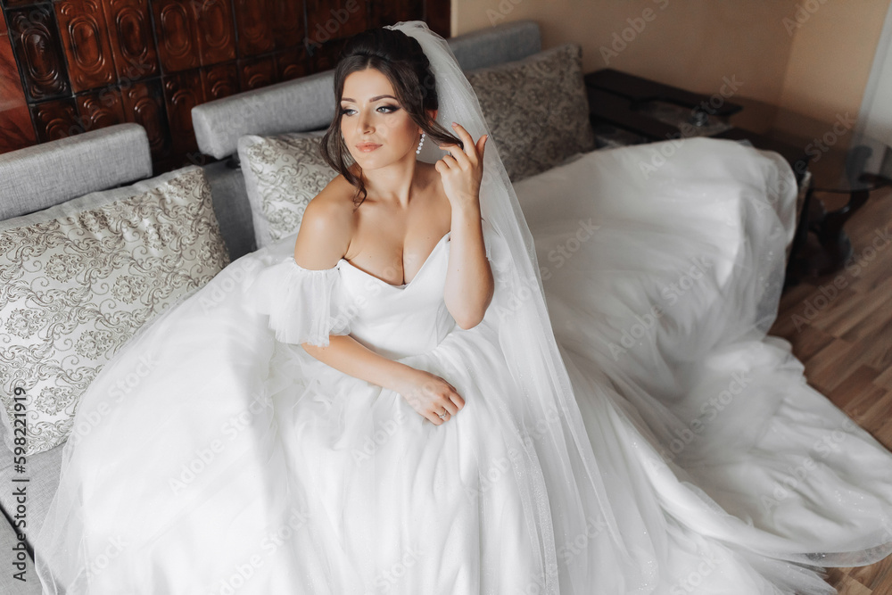 A brunette bride in a white dress poses while sitting on a gray sofa and holding her curls. Gorgeous makeup. Open shoulders. Beautiful hands. Long veil. Morning of the bride. Portrait