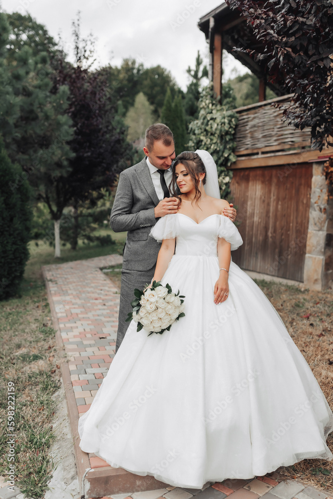 Portrait of the bride and groom in nature. The groom hugs the bride from behind, the bride in a white dress holds a bouquet of white roses and looks to the side. Beautiful makeup and hair.