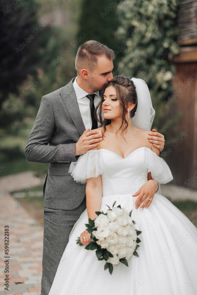 Portrait of the bride and groom in nature. The groom hugs the bride from behind and kisses her, the bride in a white dress holds a bouquet of white roses and looks away. Beautiful makeup and hair. Portrait of the bride and groom in nature. The groom hugs the bride from behind and kisses her, the bride in a white dress holds a bouquet of white roses and looks away. Beautiful makeup and hair.