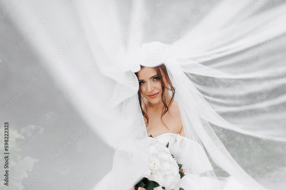Curly brunette bride in a white dress, covered with a veil, poses for the camera with a bouquet of roses. Portrait of the bride. Beautiful makeup and hair. Wedding in nature Curly brunette bride in a white dress, covered with a veil, poses for the camera with a bouquet of roses. Portrait of the bride. Beautiful makeup and hair. Wedding in nature