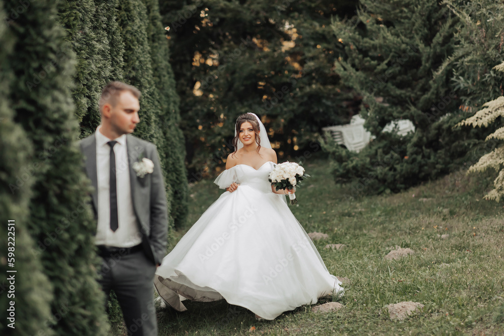 portrait of the bride and groom in nature. The groom in a gray suit is in the foreground, the bride is walking behind him, smiling in a white voluminous dress, holding a bouquet. Stylish groom
