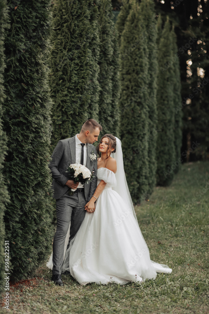 portrait of the bride and groom in nature. The groom is holding a bouquet, leaning against the bride, who is hugging him from behind, against the background of conifers. Stylish groom