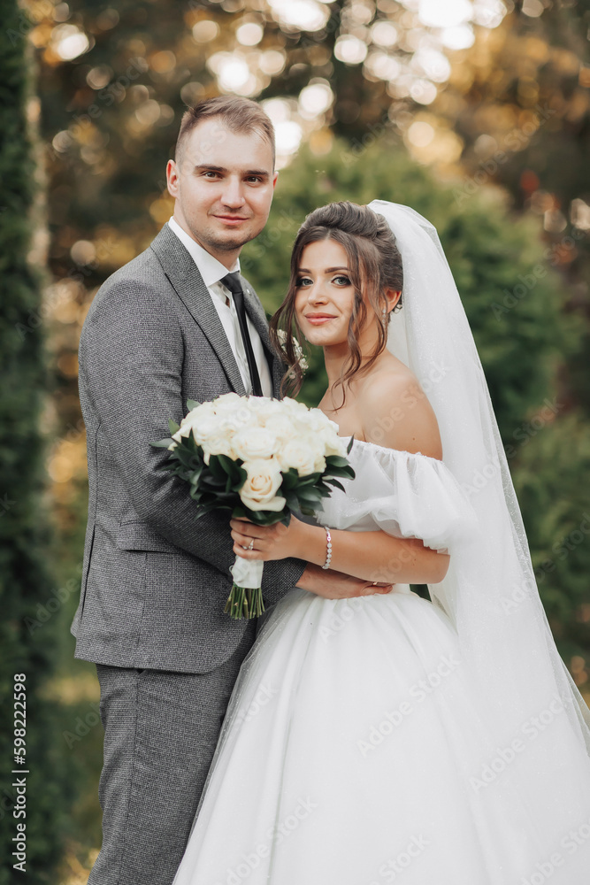 Portrait of the bride and groom in nature. Cropped photo. The bride and groom pose, hugging and smiling, against the background of conifers. The bride in a long dress with a bouquet of roses. Portrait of the bride and groom in nature. Cropped photo. The bride and groom pose, hugging and smiling, against the background of conifers. The bride in a long dress with a bouquet of roses.