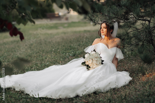 portrait of the bride in nature. A brunette bride in a white voluminous dress is sitting, posing near a coniferous tree, holding a bouquet of white roses. Beautiful hair and makeup. Warm light