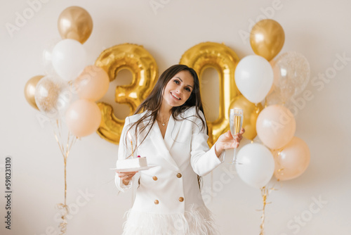 Young caucasian woman holding a cake with a candle and a glass of champagne in honor of the thirtieth birthday in a stylish white dress with feathers