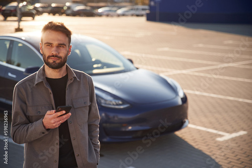 With smartphone. Man is standing near his electric car outdoors © standret