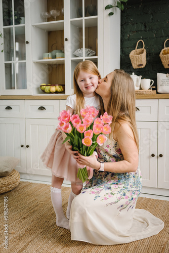 The daughter congratulates mother and gives present, bouquet of tulip flowers, in kitchen at home. Mother's Day concept. Mom kiss child girl smiling. Greeting card. International Womens Day.