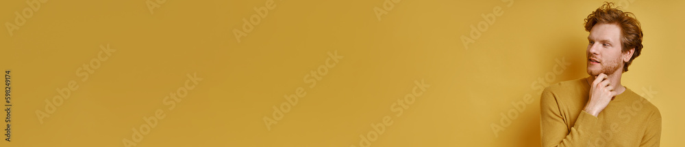 Thoughtful redhead man holding hand on chin while standing against yellow background