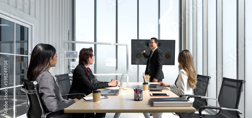 Asian senior manager in black suit giving presentation with large digital monitor. Business executive team meeting in modern office with laptop computer, tablet, mobile phone and coffee on table.