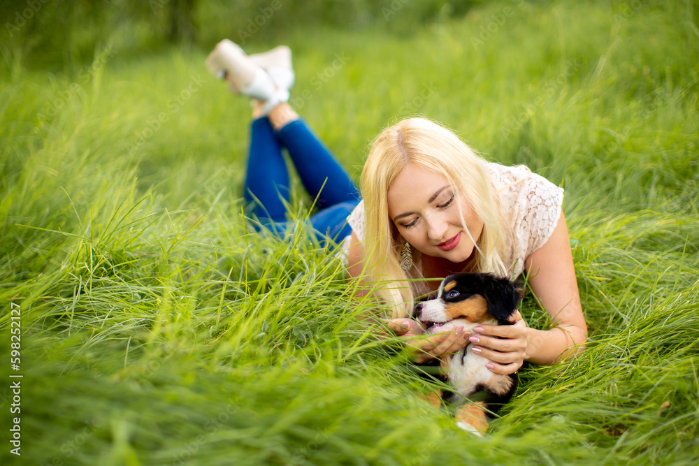 Fototapeta premium A beautiful girl with a dog sits in the park on the grass. Love to the animals. Home pet on a walk. Animal protection. Bernese mountain dog puppy.