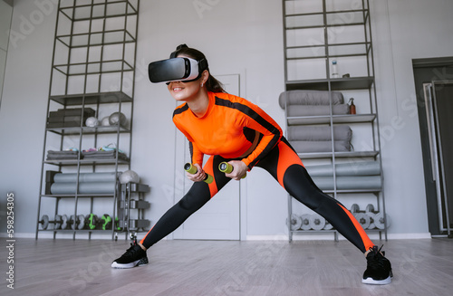 Fit brunette hispanic young woman in orange sportswear training with dumbbells using 3D headset at sport club. Excited American girl in virtual reality googles at fitness club. Healthy people. © Iona