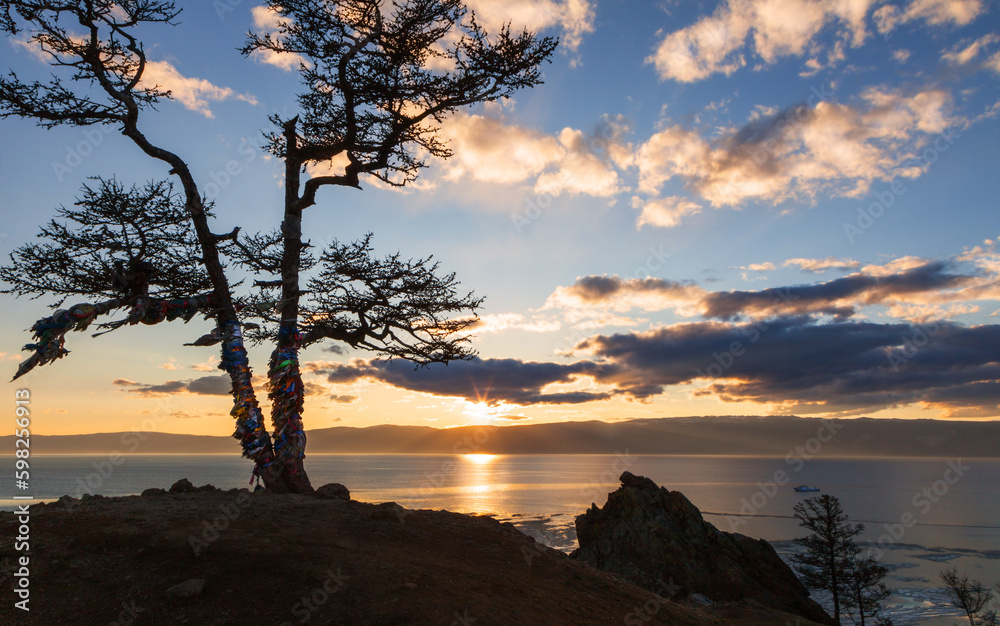 Scenic spring sunset at Baikal Lake. View of Small Sea from coast of Olkhon Island. Silhouette of famous Wish Tree with colorful ribbons of tourists. Natural background. Travel and outdoor recreation Scenic spring sunset at Baikal Lake. View of Small Sea from coast of Olkhon Island. Silhouette of famous Wish Tree with colorful ribbons of tourists. Natural background. Travel and outdoor recreation