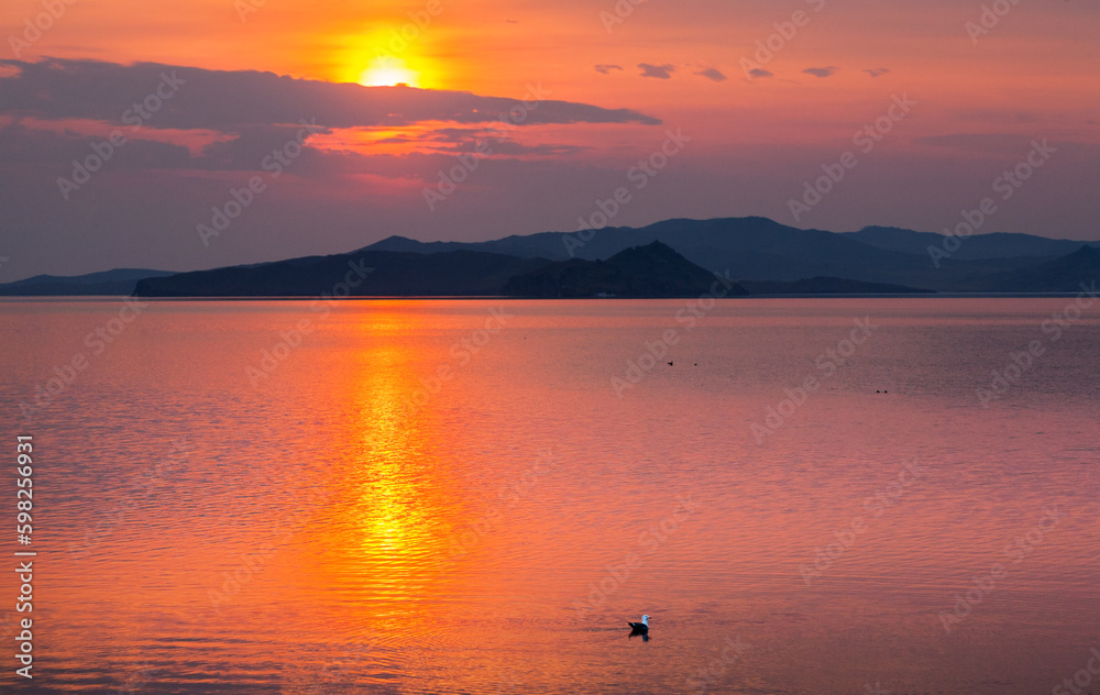 A picturesque colorful dawn is reflected in water of Baikal Lake in early spring morning. A seagull sways on waves of Small Sea. Beautiful seascape. Natural background. Travel and outdoors recreation