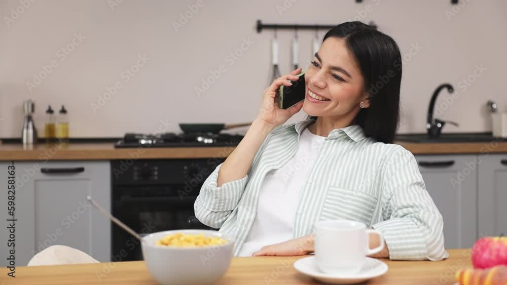 Young happy woman wear casual clothes shirt hold in hand use mobile cell phone talk speak conducting pleasant conversation sit at table in light kitchen at home alone. Lifestyle cooking food concept Young happy woman wear casual clothes shirt hold in hand use mobile cell phone talk speak conducting pleasant conversation sit at table in light kitchen at home alone. Lifestyle cooking food concept