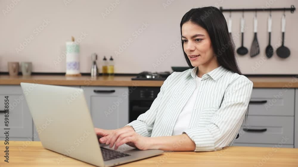 Young calm happy housewife smart IT user woman wear casual clothes shirt use work on laptop pc computer typing sit at table in light kitchen office study at home alone. Lifestyle cooking food concept Young calm happy housewife smart IT user woman wear casual clothes shirt use work on laptop pc computer typing sit at table in light kitchen office study at home alone. Lifestyle cooking food concept