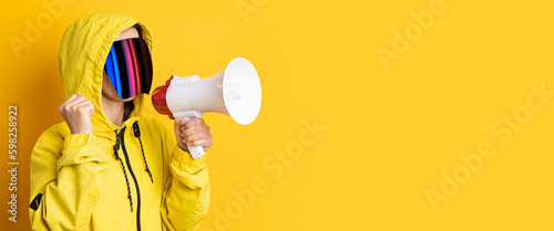 Young woman in cyberpunk glasses with a megaphone in a yellow jacket on a yellow background. Banner.