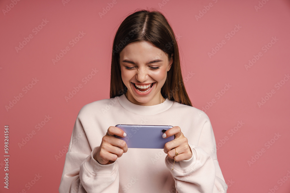 Excited woman playing online game on smartphone isolated over pink background Excited woman playing online game on smartphone isolated over pink background