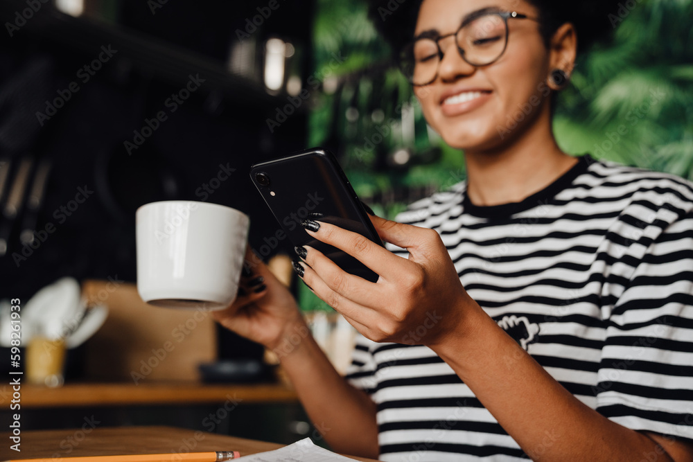 African woman using smartphone and drinking coffee while sitting in kitchen African woman using smartphone and drinking coffee while sitting in kitchen