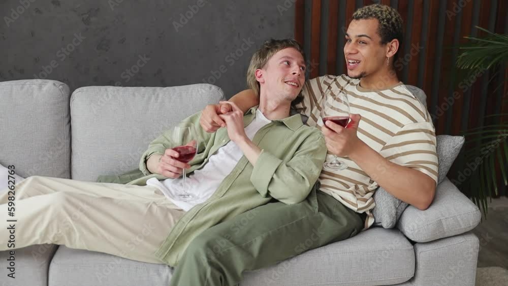 Young happy couple two gay men wear casual clothes drink wine talk speak lay on sofa couch stay at home flat rest relax spend free spare time in living room. Pride day june month love lgbtq concept Young happy couple two gay men wear casual clothes drink wine talk speak lay on sofa couch stay at home flat rest relax spend free spare time in living room. Pride day june month love lgbtq concept