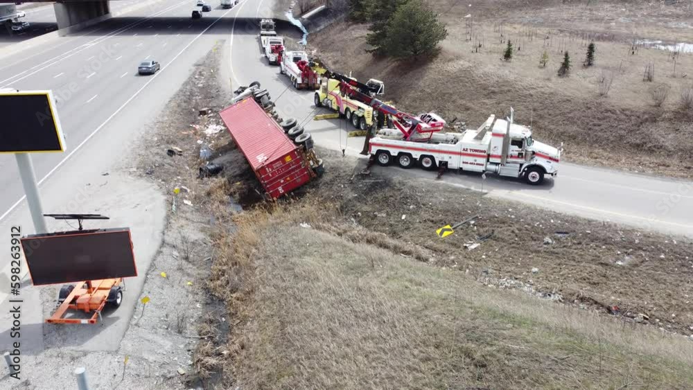 Large red semi truck flipped over at highway on-ramp, tow trucks attempt to pull it back