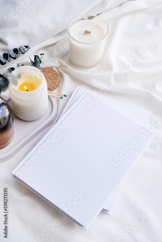 Soy wax aroma candle in white jar on bed with eucalyptus leaves. Candle and book mockup design