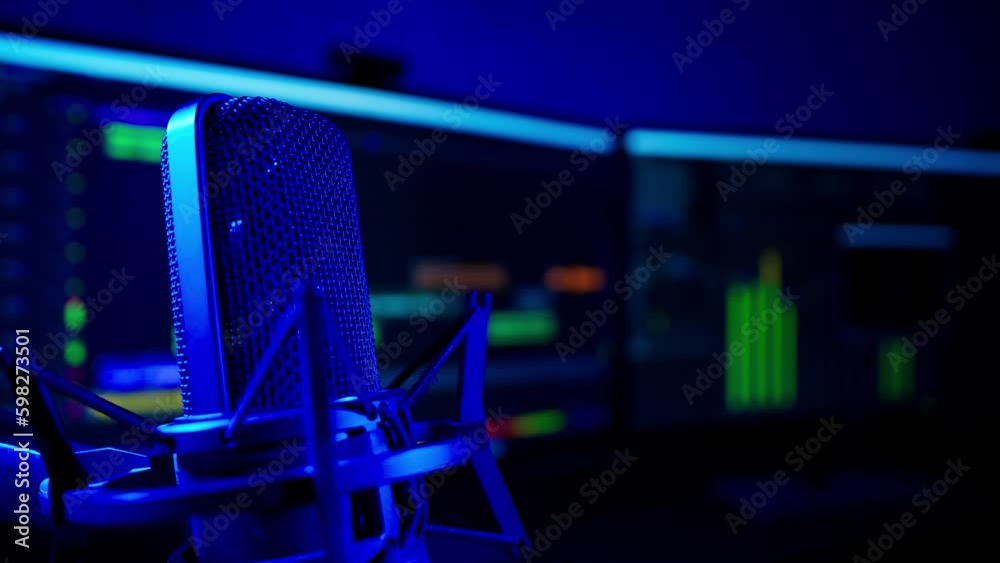 Close shot of a professional studio condenser microphone with audio editing software recording in soft-focus in the background. The sound recording studio is lit with a deep blue light.
