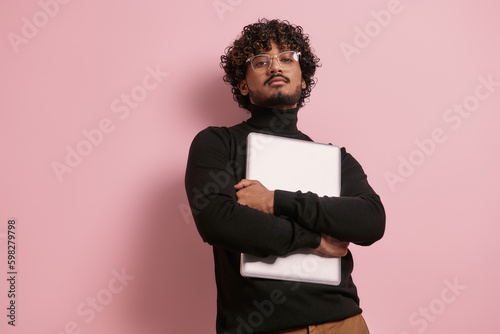 Confident young Indian man looking at camera while carrying laptop against pink background Generative AI