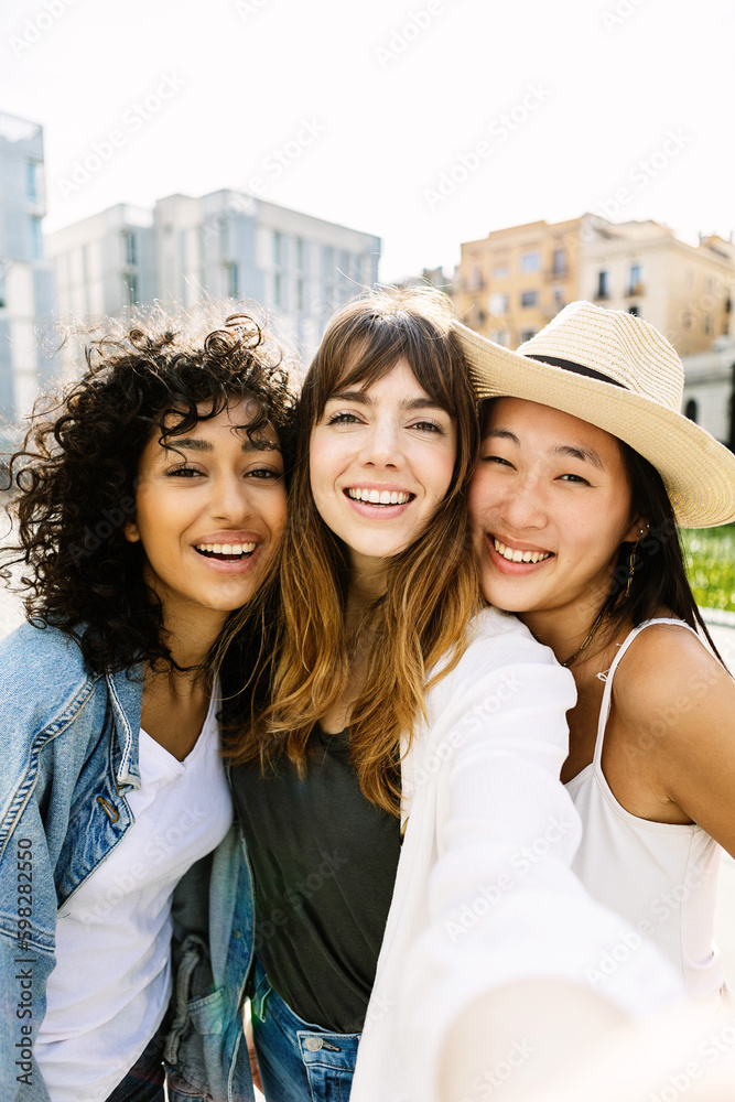 Young group of three diverse women taking selfie portrait with phone outdoors. Joyful millennial female friends enjoying day off at weekend in city street. Friendship concept Generative AI - obrazy, fototapety, plakaty 