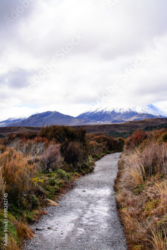 Gravel track going through desolated high land field in Central Plateau of New Zealand. Snow blanketed volcanic cone of Mount Ngauruhoe covered by low clouds. Tongariro National Park, North Island