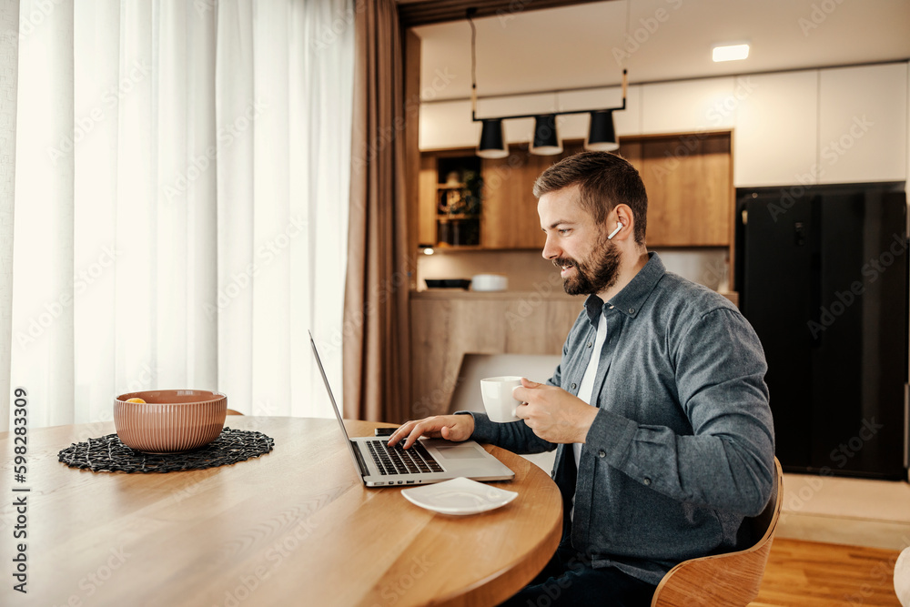 A happy entrepreneur is enjoying his coffee while working from home on a laptop. A happy entrepreneur is enjoying his coffee while working from home on a laptop.