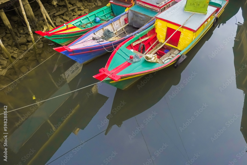 Obraz premium Many small passenger ships is at anchor between wooden buildings in shallow water. Colorful fishing boats in harbor during the day. Small engine boats.