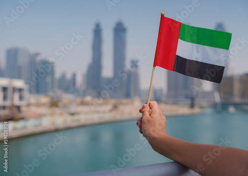 Photography Close up of a female hand holding tiny flag of UAE against Dubai skyline
