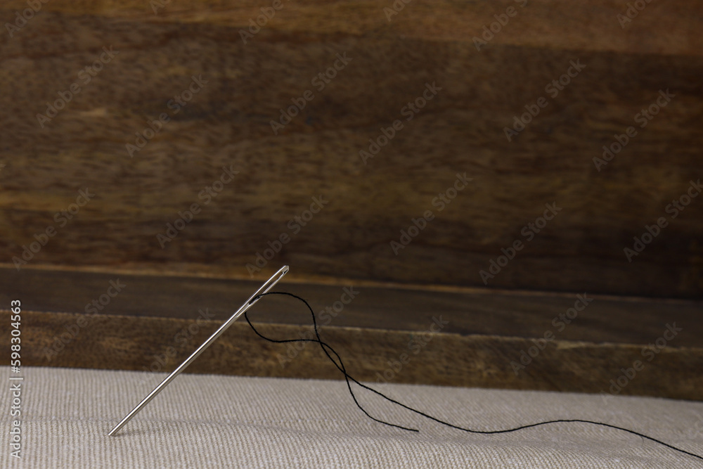 Steel Needle with black thread sticking in a natural uncolored cotton cloth in front of brown wooden background Steel Needle with black thread sticking in a natural uncolored cotton cloth in front of brown wooden background
