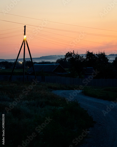 Village landscape. Sunset, dawn, pink sky, dusk, small houses, electric pole with wires in the foreground, road, trees and bushes, lantern on a pole