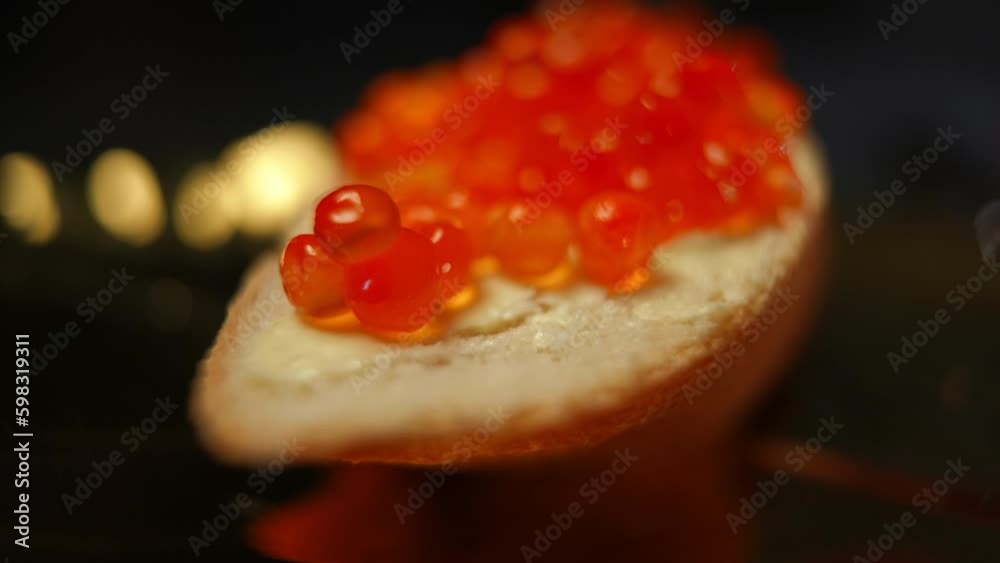 Close-up placing round red salmon roe on butter with bread in slow motion. Closeup delicious tasty sandwich in darkness indoors