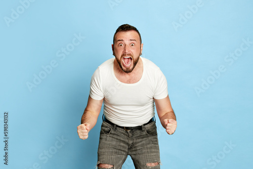 Portrait of bearded man in white t-shirt posing with excited,positive, winning emotions against blue studio background. Concept of human emotions, lifestyle, facial expression, success. Ad