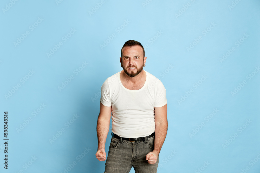 Portrait of bearded man in casual white t-shirt showing anger and irritation, holding fists clenched against blue studio background. Concept of human emotions, lifestyle, facial expression. Ad Portrait of bearded man in casual white t-shirt showing anger and irritation, holding fists clenched against blue studio background. Concept of human emotions, lifestyle, facial expression. Ad