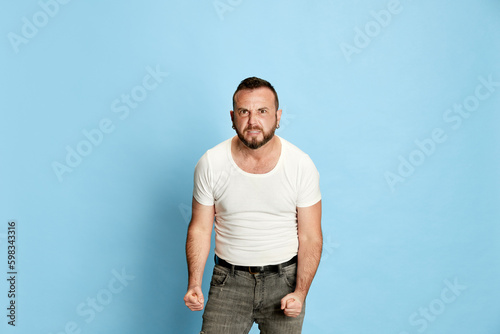 Portrait of bearded man in casual white t-shirt showing anger and irritation, holding fists clenched against blue studio background. Concept of human emotions, lifestyle, facial expression. Ad