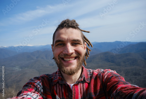 Concept of healthy active lifestyle. Young travel blogger with dreadlocks went hiking in mountains. Man enjoys views of nature takes video or selfie photo on phone or camera. Wide-angle view portrait.