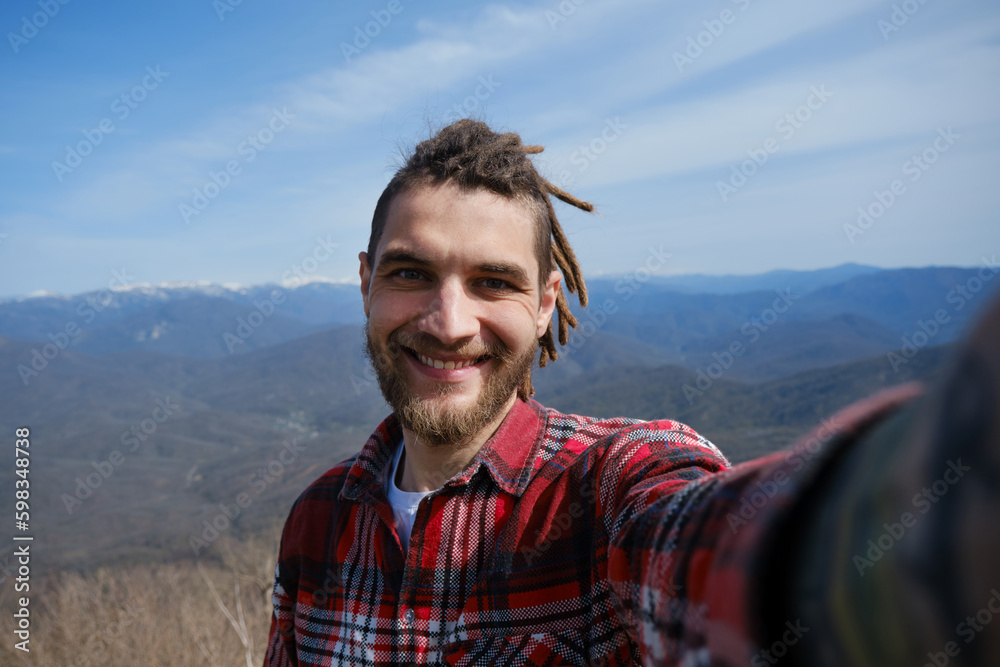 Concept of healthy active lifestyle. Young travel blogger with dreadlocks went hiking in mountains. Man enjoys views of nature takes video or selfie photo on phone or camera. Wide-angle view portrait.