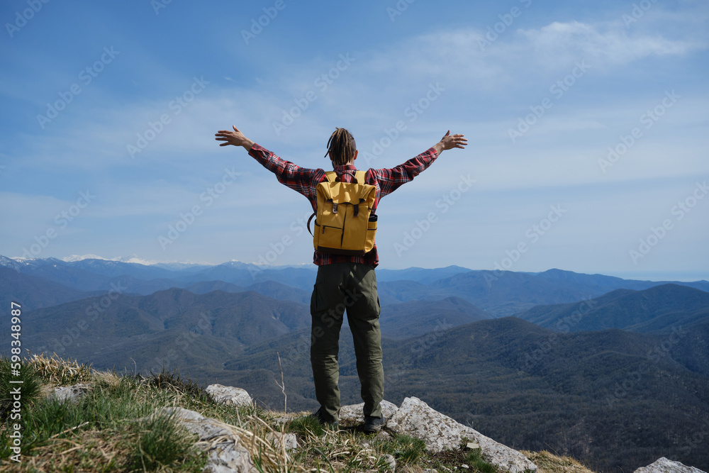 Young guy with dreadlocks went hiking in mountains. Man with yellow backpack stands on top of hill and spreads arms in different directions. Rear view. Concept of travel and healthy, active lifestyle. Young guy with dreadlocks went hiking in mountains. Man with yellow backpack stands on top of hill and spreads arms in different directions. Rear view. Concept of travel and healthy, active lifestyle.