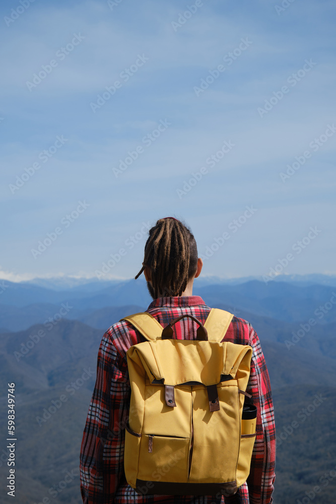 Concept of travel and healthy, active lifestyle. Man with yellow backpack stands on top of hill and enjoys views of nature Caucasus. Rear view. Young guy with dreadlocks went hiking in mountains.