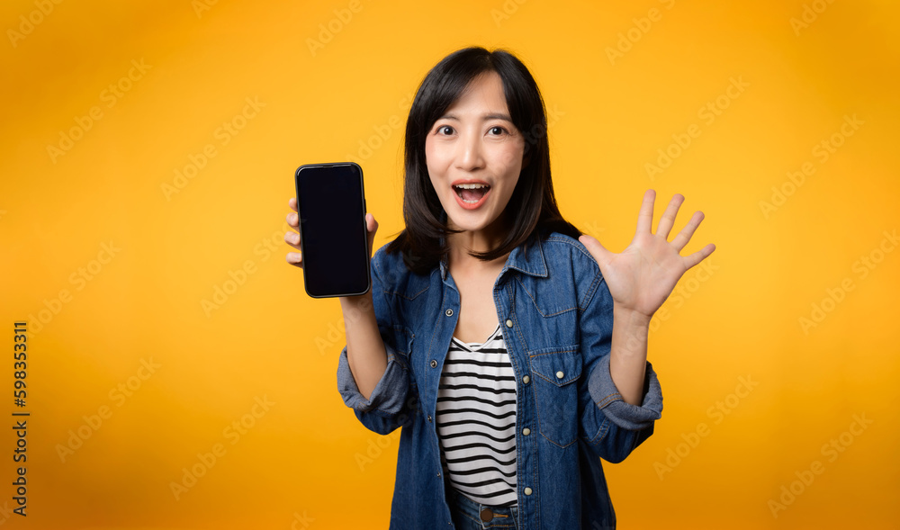 Portrait beautiful young asian woman happy smile dressed in denim jacket showing smartphone screen isolate on yellow studio background. New smartphone application concept