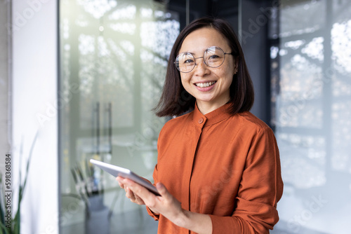 Portrait of a young successful Asian female programmer inside office, worker in glasses smiling and looking at the camera, a businesswoman holds a tablet computer in hands and uses it to test online . © Liubomir