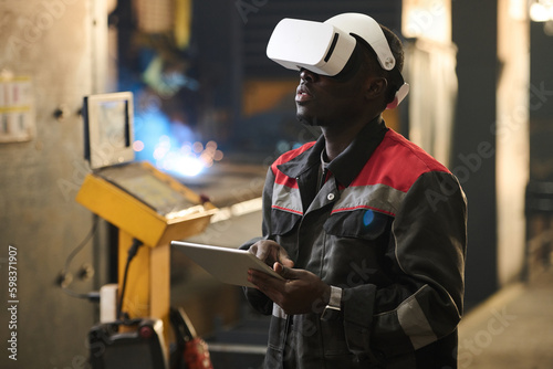 Young African American worker of factory in vr headset using tablet while operating industrial machine during technical production process