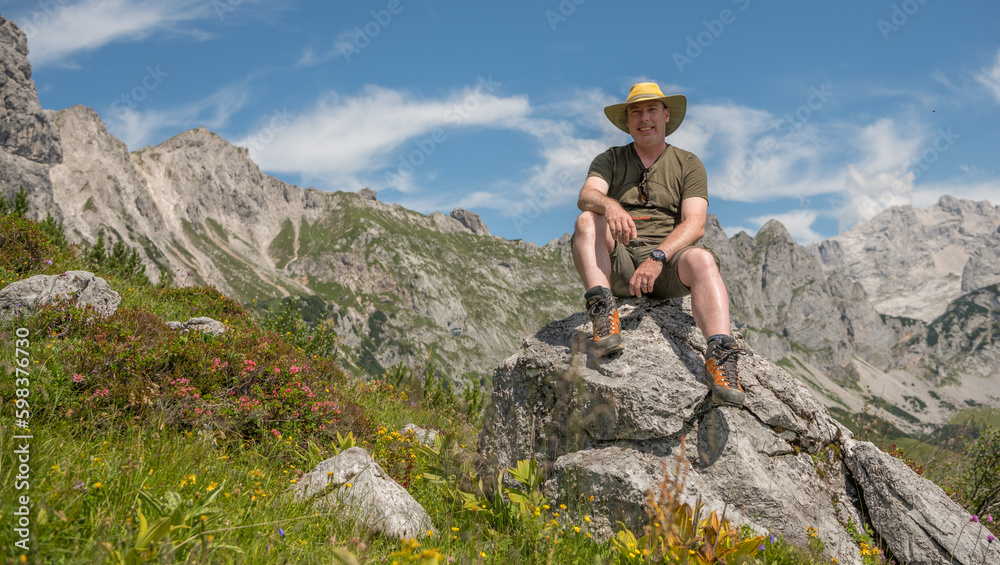 Caucasian hiker sitting and resting on a rock with beautiful Austrian mountains in the background. the hiker is wearing a short, T-shirt and hat. Austria, summer, hiking, sunny. happy, happiness.