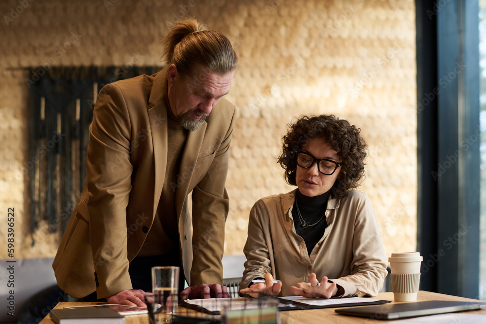 Fototapeta premium Confident businesswoman explaining data in document to mature male colleague standing by her and bending over table during discussion